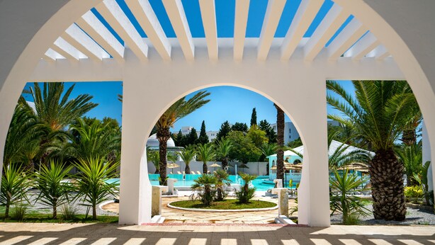 Zona de piscina en un resort, con vista despejada a la piscina, rodeada de palmeras y una estructura arqueada blanca.