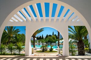 Un espace piscine aménagé dans un complexe hôtelier, offrant une vue dégagée sur la piscine, entouré de palmiers et agrémenté d’une structure en forme d’arche blanche.