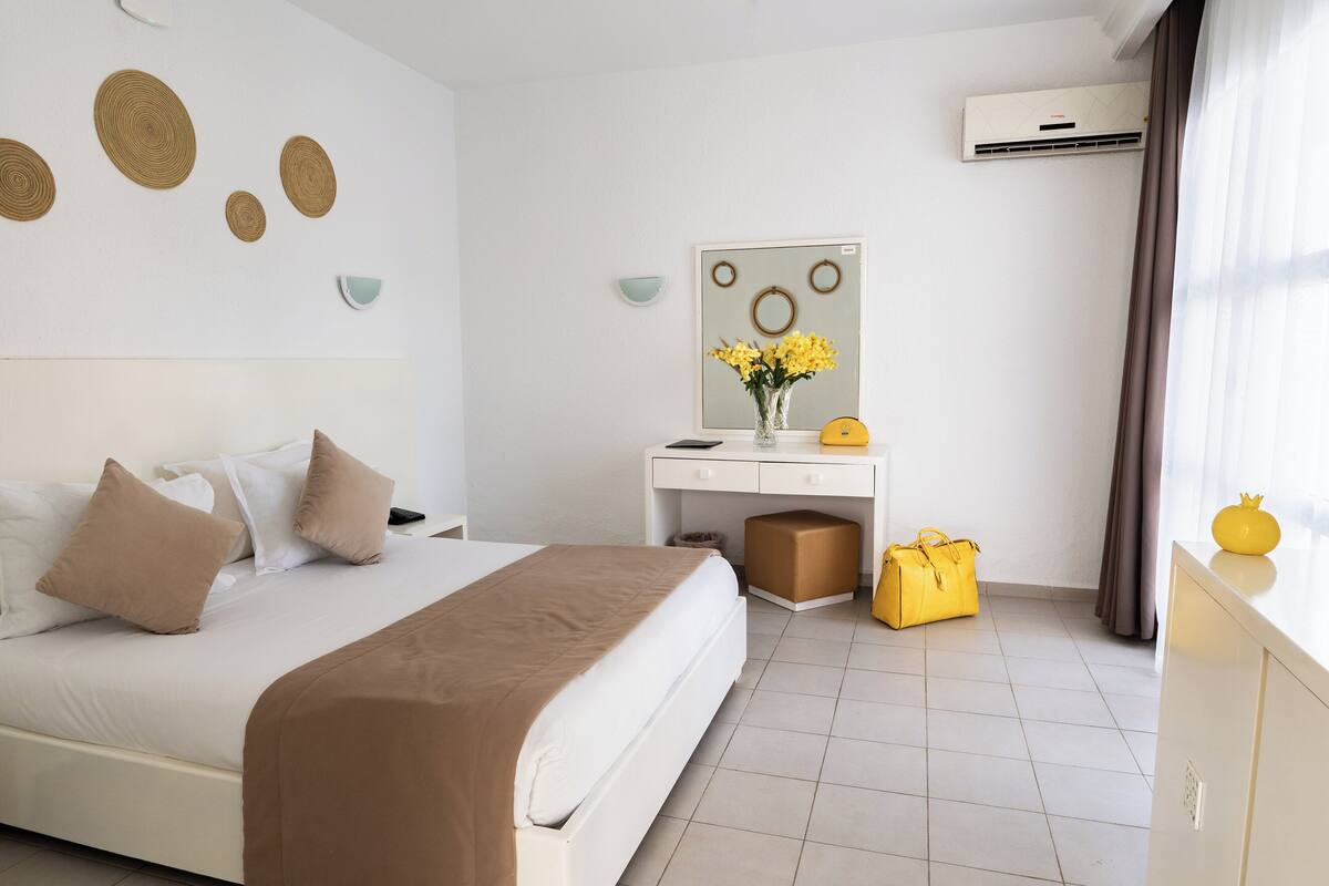 A hotel room with a bed, a desk, a chair, a potted plant, and a window with curtains.