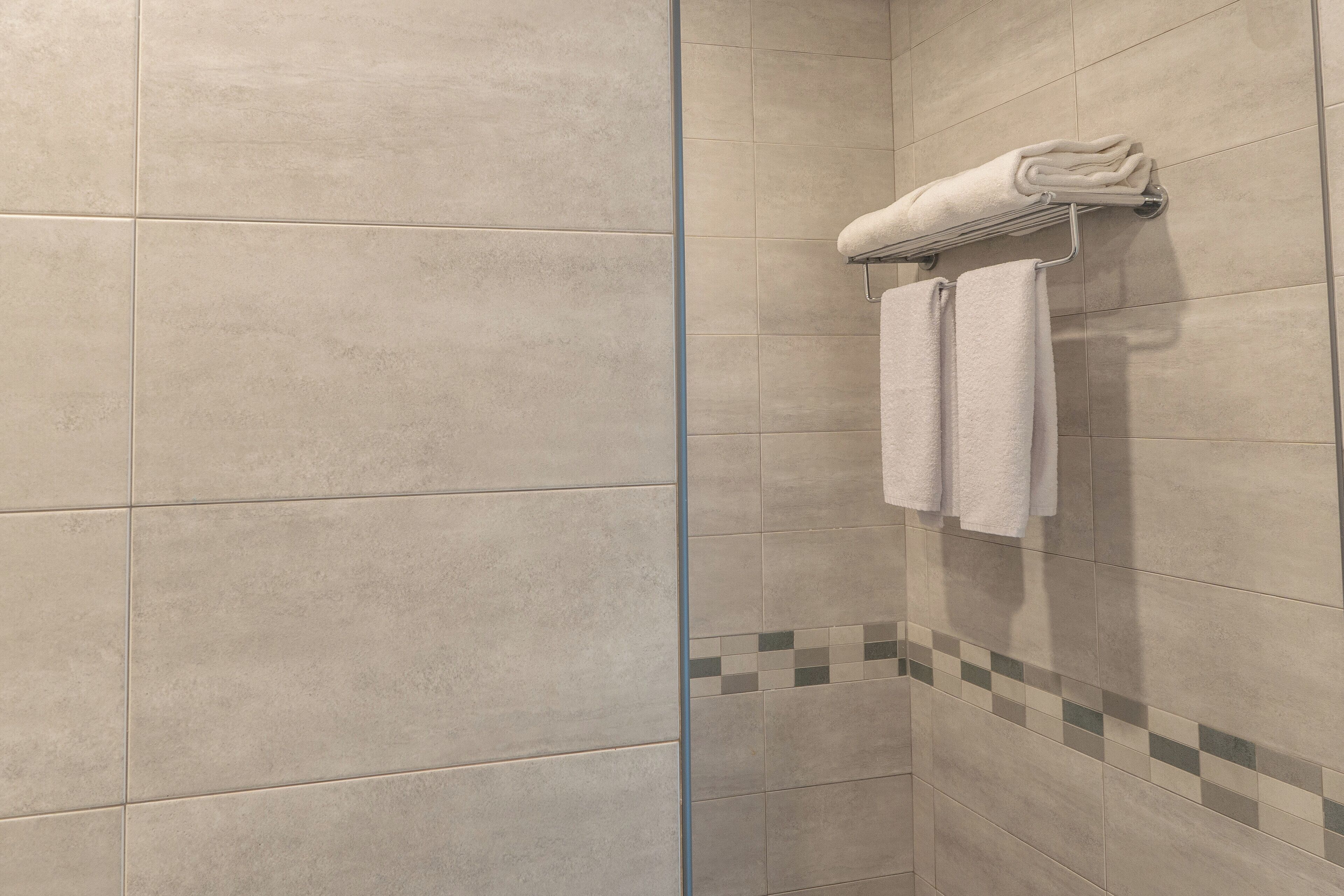 Un baño con baldosas color beige, una puerta de vidrio para la ducha y un perchero con toallas dobladas.