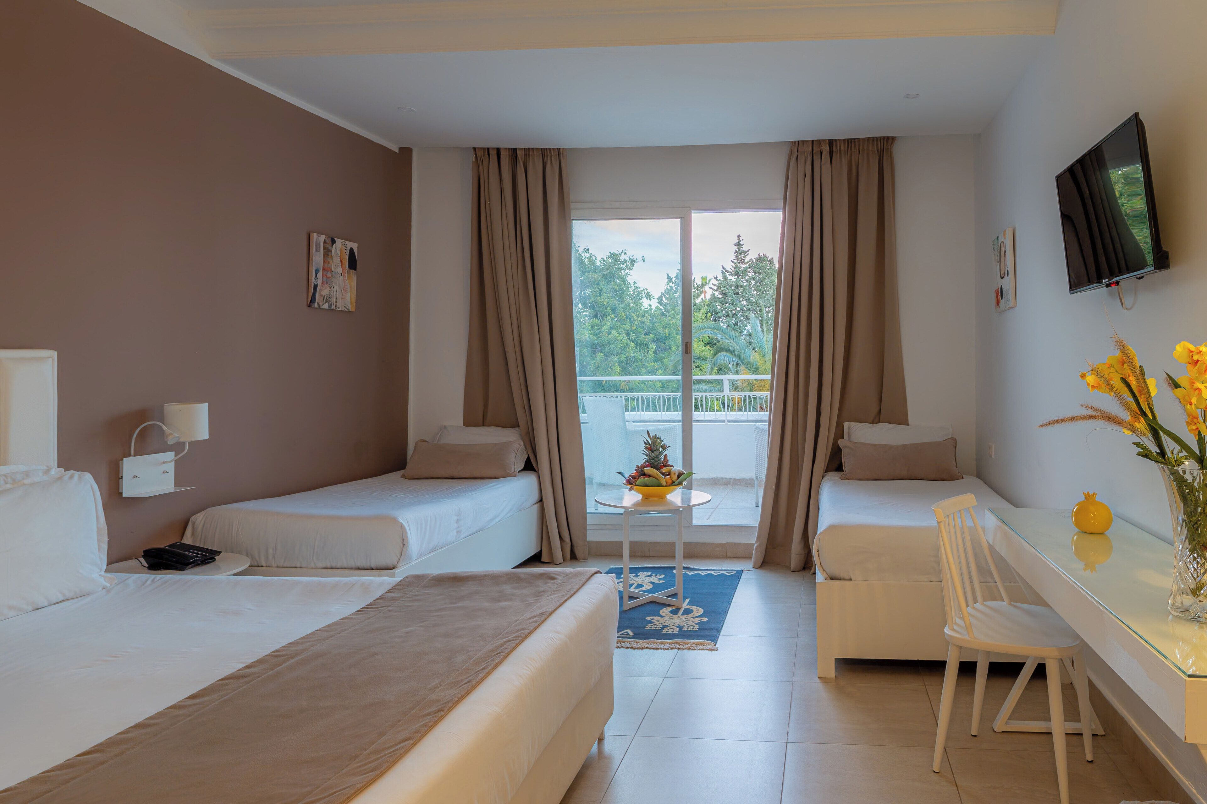 A hotel room with two beds, a desk, a chair, a TV, and a balcony with a view of greenery.