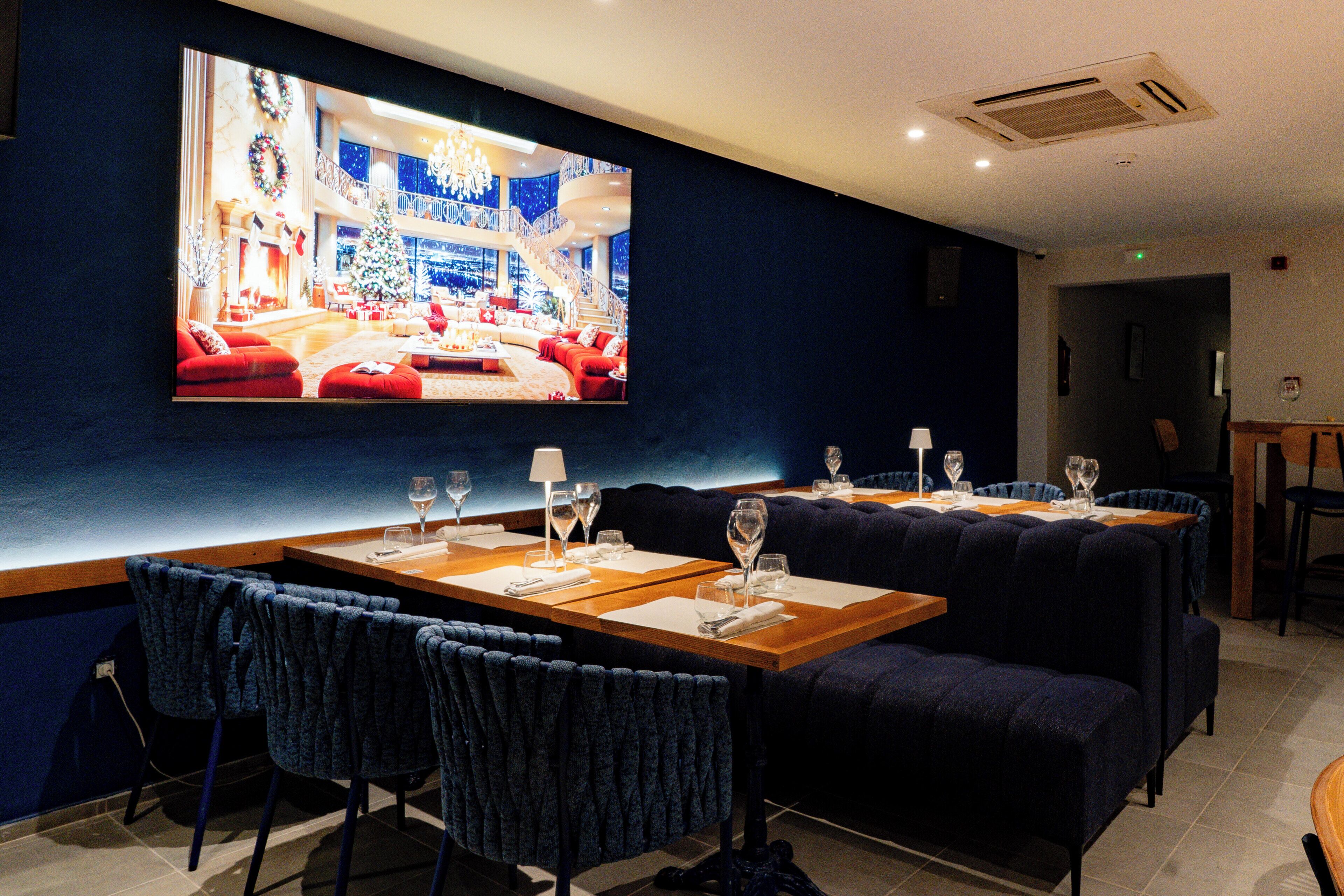Un comedor moderno con una tele grande que muestra una escena festiva, asientos tapizados en azul oscuro y una mesa de madera con copas de vino.