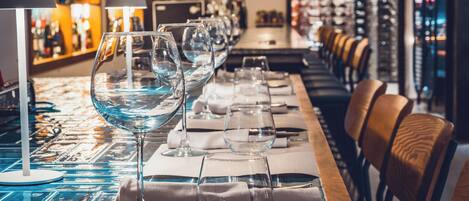 Una zona de bar con copas de vino y servilletas ordenadas sobre una mesa, y una barra con sillas al fondo.
