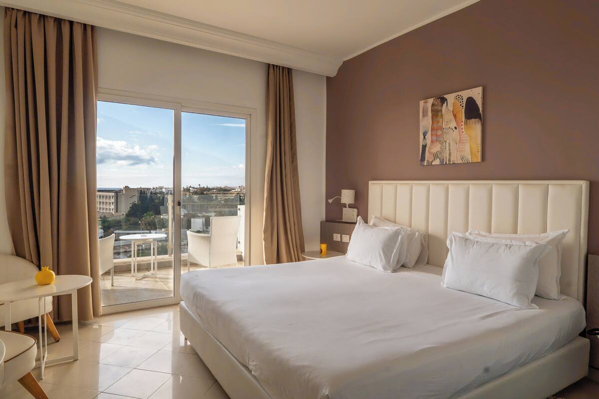 A hotel room with a large bed, a balcony with a view, and a dining area.