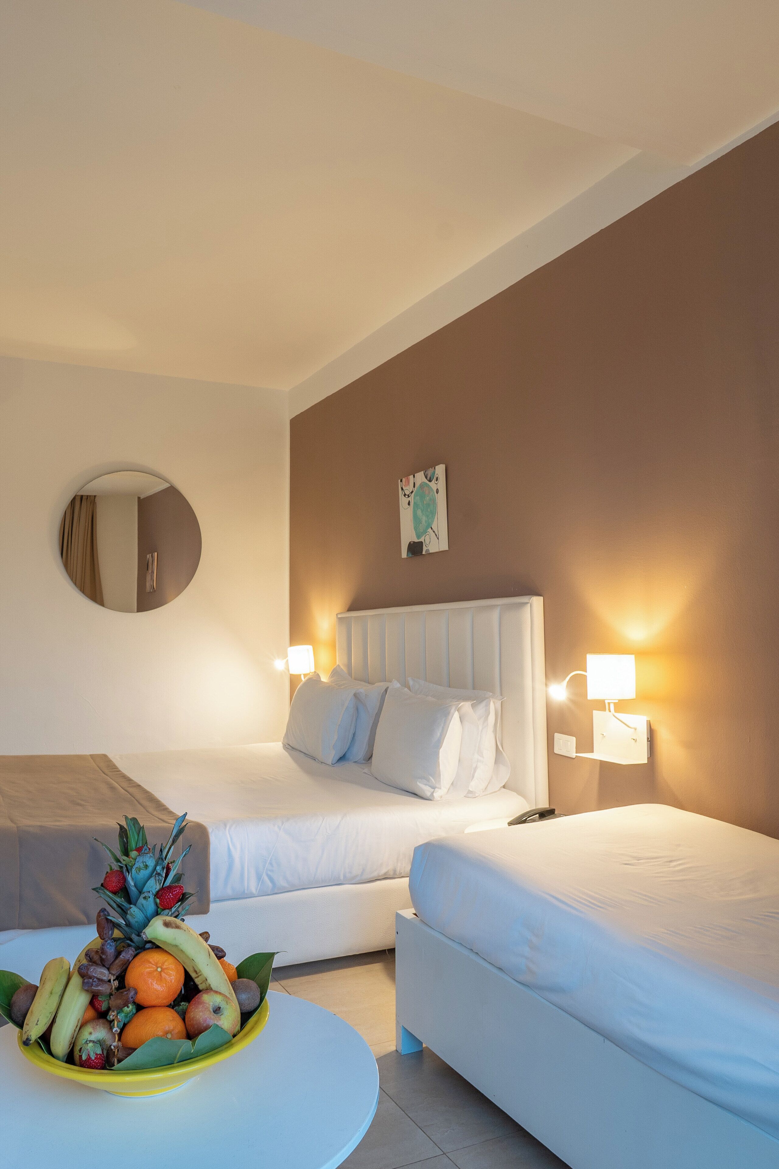 A hotel room with two beds, a round mirror, a fruit bowl on a small table, and wall-mounted lamps.