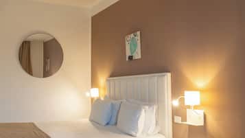 A hotel room with two beds, a round mirror, a fruit bowl on a small table, and wall-mounted lamps.