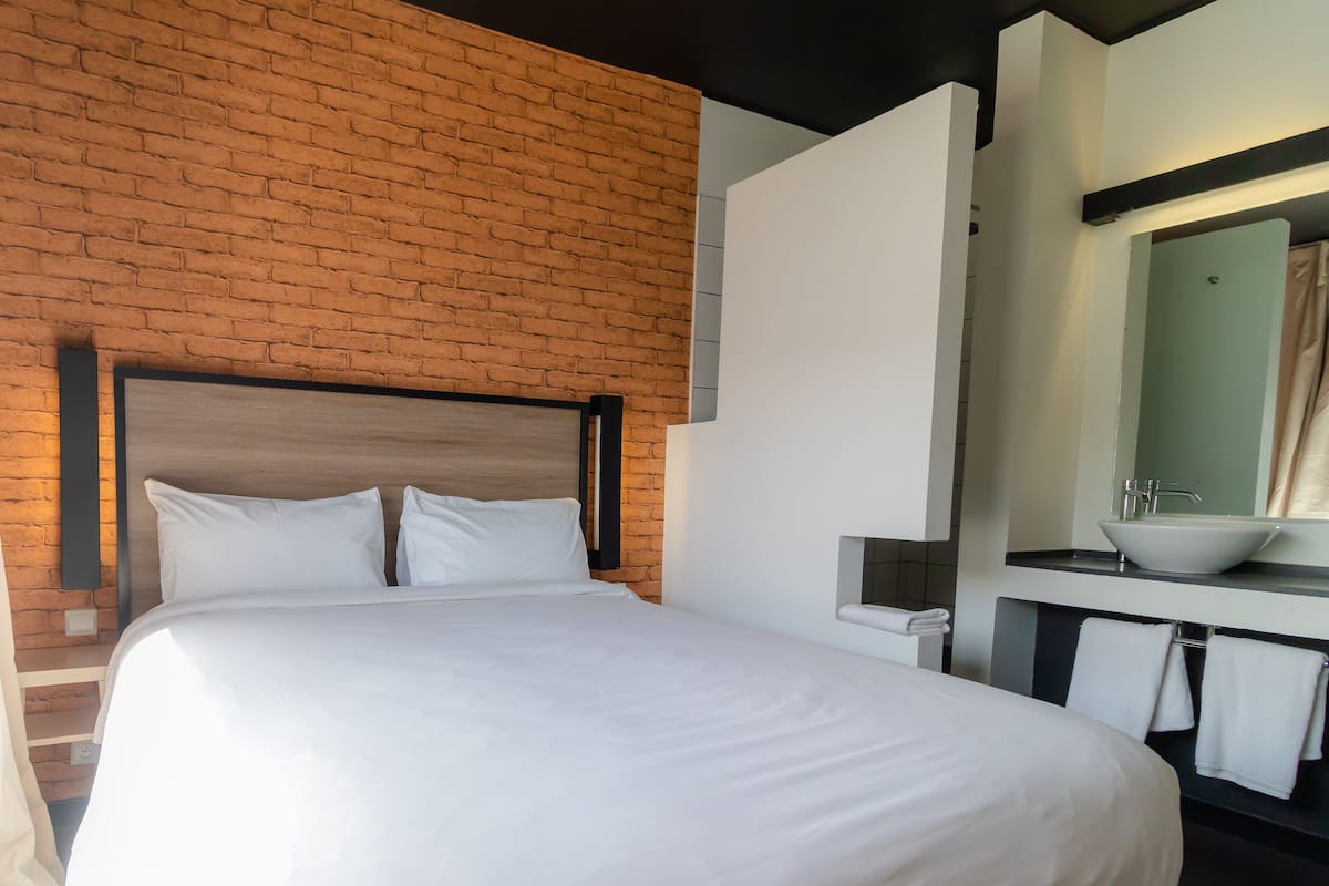 A hotel room with a brick wall, a bed with white bedding, a bedside table, and a bathroom with a sink and mirror.
