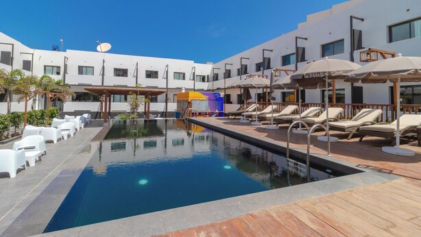 Un área de piscina con sillones de descanso y sombrillas, rodeada por un edificio moderno con numerosas ventanas y un cielo azul despejado.