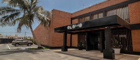Um edifício com design moderno, fachada de tijolos e marquise preta com um logotipo. Há uma palmeira e um carro estacionado em primeiro plano.
