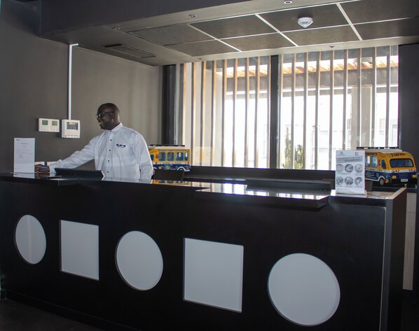 A reception desk with a man standing behind it, a window with vertical blinds, and a toy bus model on the counter.