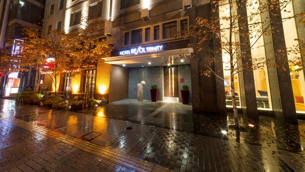 A hotel entrance with a sign reading "HOTEL RESOL TRINITY," a wet pavement, and trees with autumn leaves. - Hotel Resol Trinity Sapporo (Sapporo)