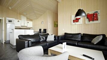 A modern living room with a black leather sofa, a white coffee table, and a grey armchair. There is a kitchen area with white cabinets and a refrigerator. A black pendant light hangs above the sofa, and a red abstract painting is on the wall.