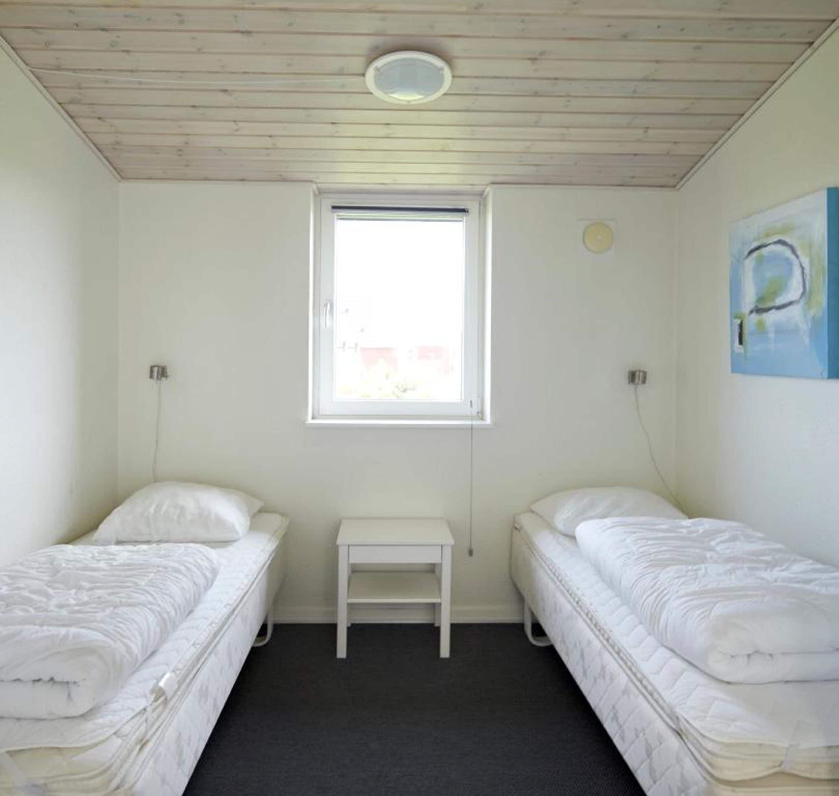A small, clean room with two single beds, a small table, and a window providing natural light.