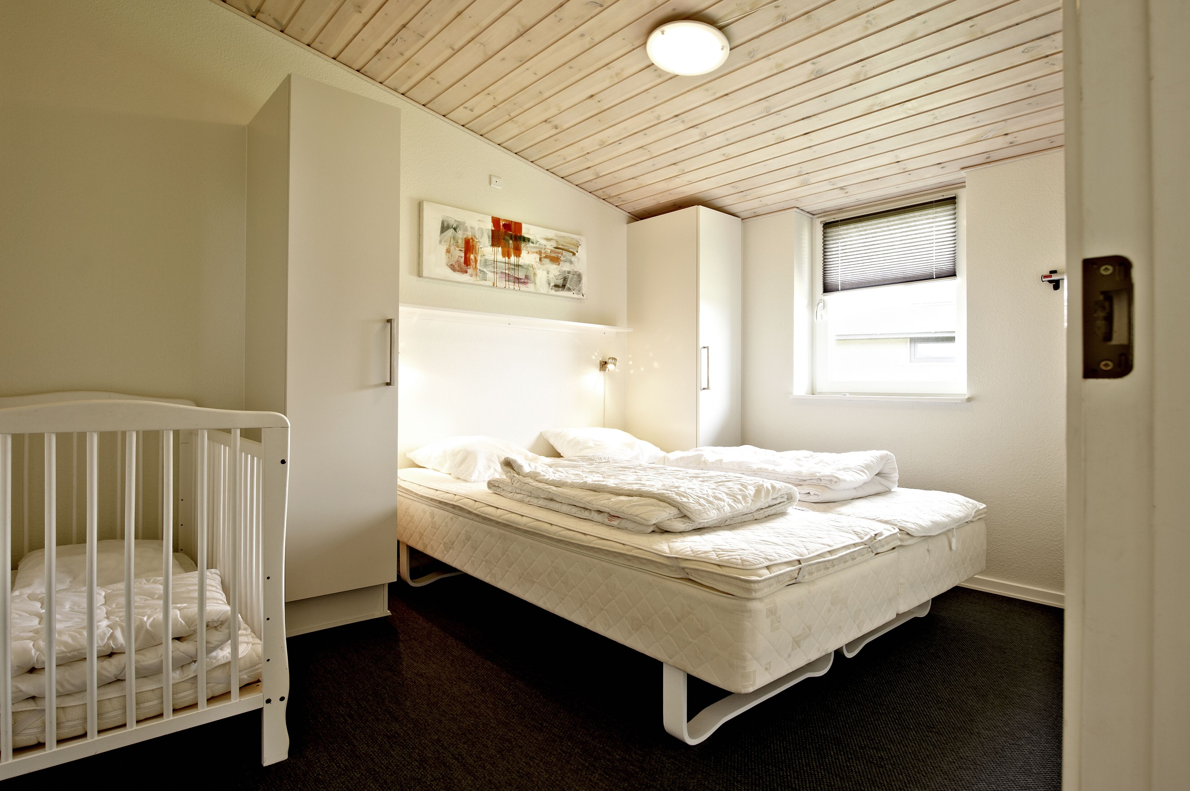 Una habitación pequeña y limpia con una cuna, una cama individual, una ventana con persianas e iluminación en el techo.