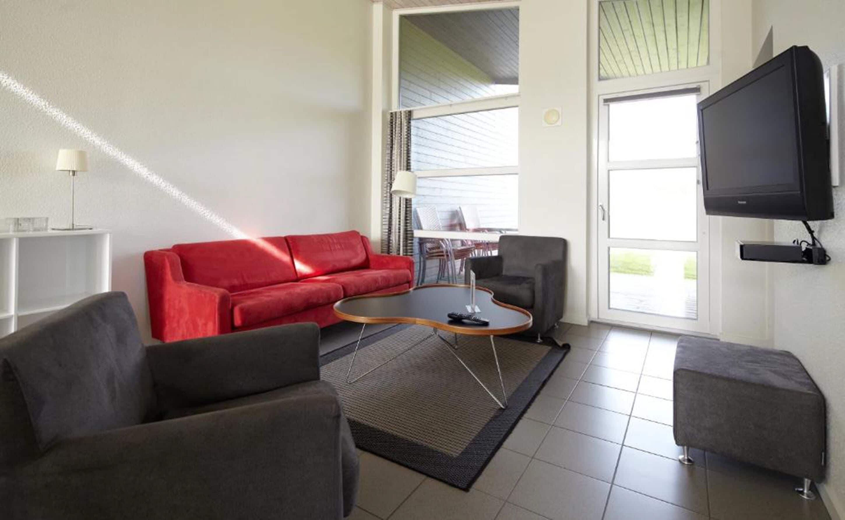 En moderne stue med en rød sofa, grå lænestole, et sofabord og et fladskærms-tv monteret på væggen.