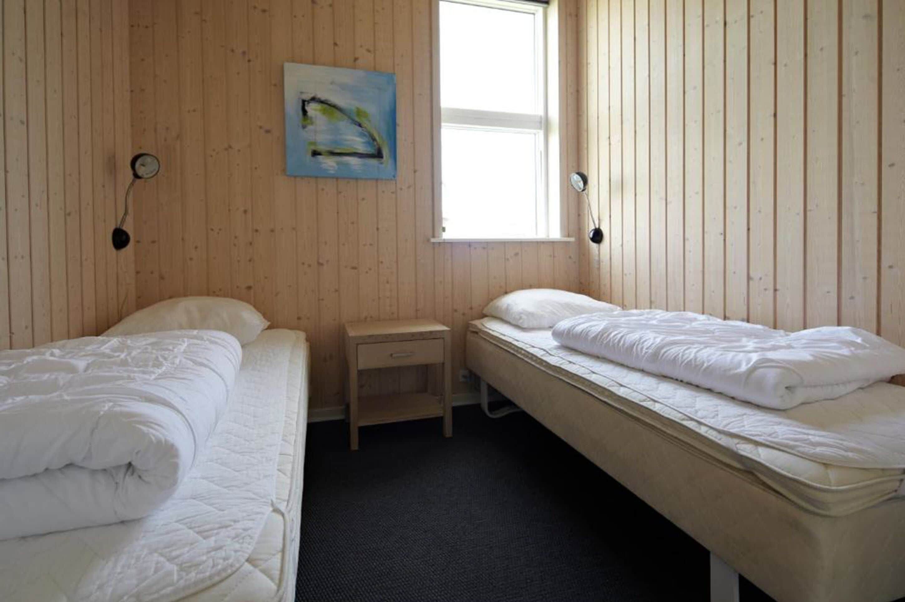 Ett rum med två sängar, ett litet träbord vid sängen, en tavla på väggen och ett fönster som släpper in naturligt ljus.