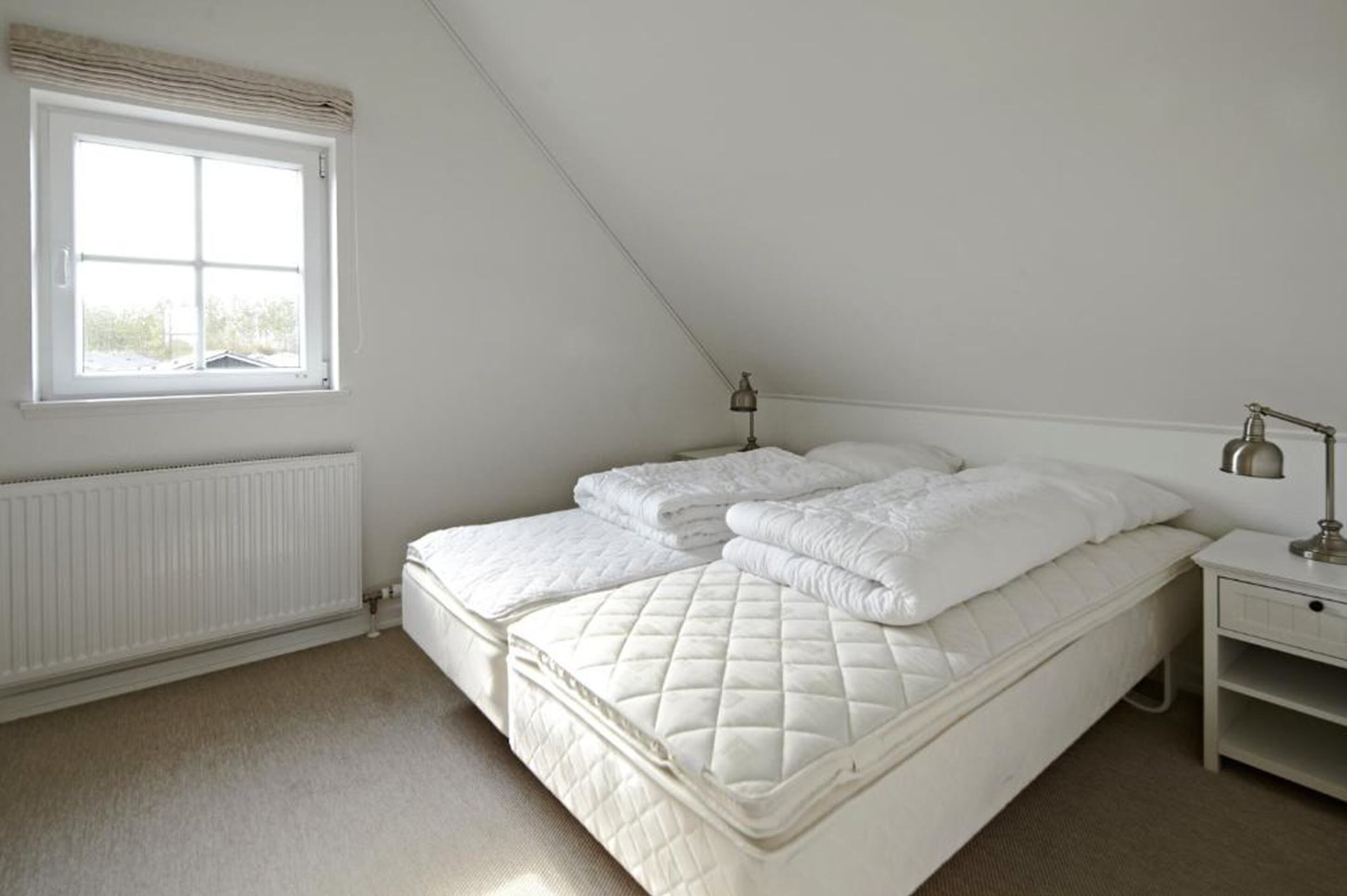 A bedroom with a white bed, a nightstand, a lamp, and a window with blinds.