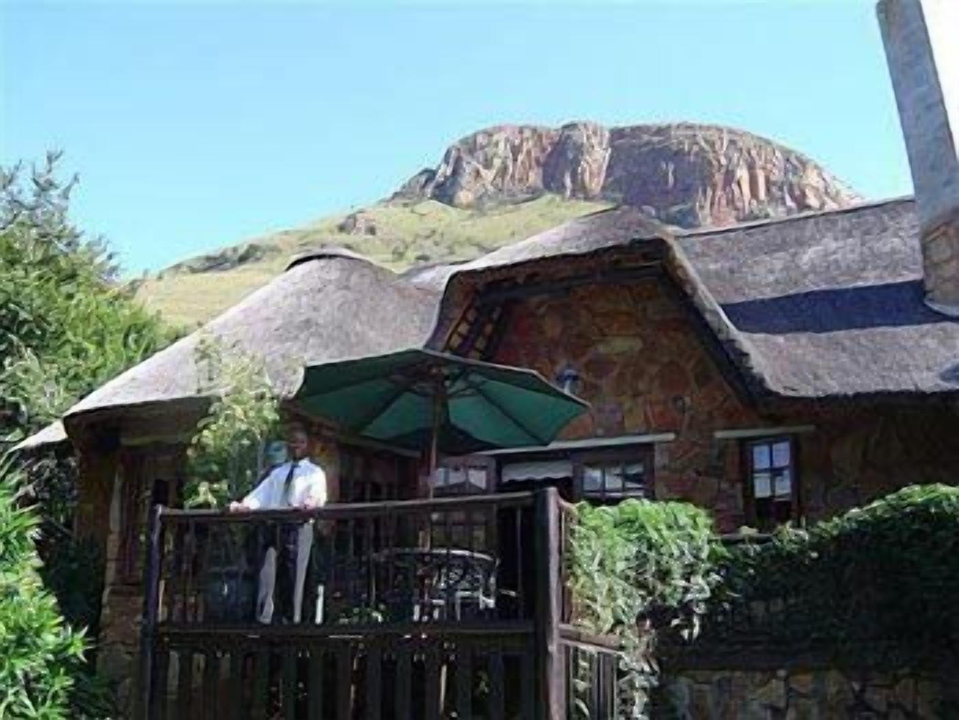 Un bâtiment rustique avec un toit de chaume, un balcon agrémenté d’une table et de chaises, et une vue sur une montagne en arrière-plan.