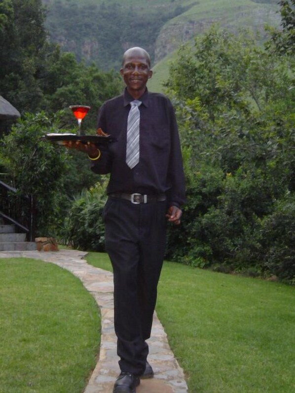 Un hombre con camisa y corbata oscura camina por un sendero de piedra en un jardín exuberante, con abundante vegetación y montañas al fondo.