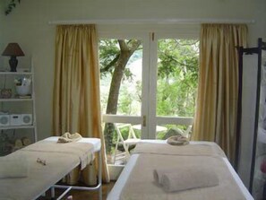A room with two beds, a window with curtains, a shelf with items, and a view of trees outside.