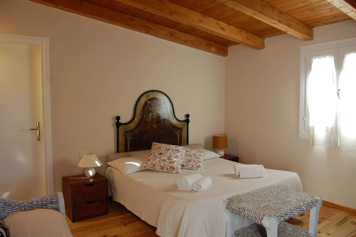 Ein Schlafzimmer mit Holzboden, einem Bett mit floralen Kissen, einem Nachttisch mit Lampe und einem Fenster mit Vorhängen.