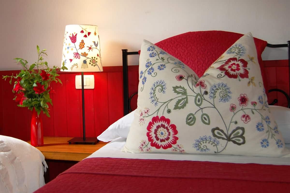 Ein Hotelzimmer mit rotem Bettüberwurf, einem blumigen Kopfkissen, einer Nachttischlampe und einer Vase mit roten Blumen.