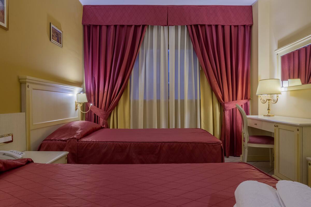 Une chambre d’hôtel avec deux lits, un bureau, une chaise et une fenêtre avec des rideaux.