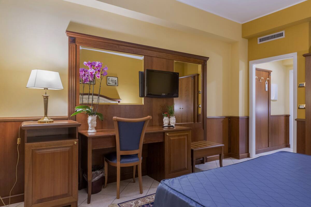 Une chambre d’hôtel avec un bureau, une chaise et un téléviseur en bois. On y trouve un lit recouvert d’une courtepointe bleue, une lampe et un vase avec des fleurs.
