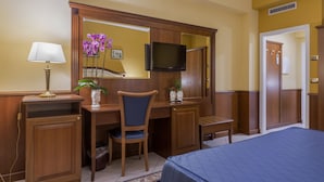 A hotel room with a wooden desk, chair, and television. There is a bed with a blue bedspread, a lamp, and a vase with flowers. - Hotel San Michele (Milazzo)
