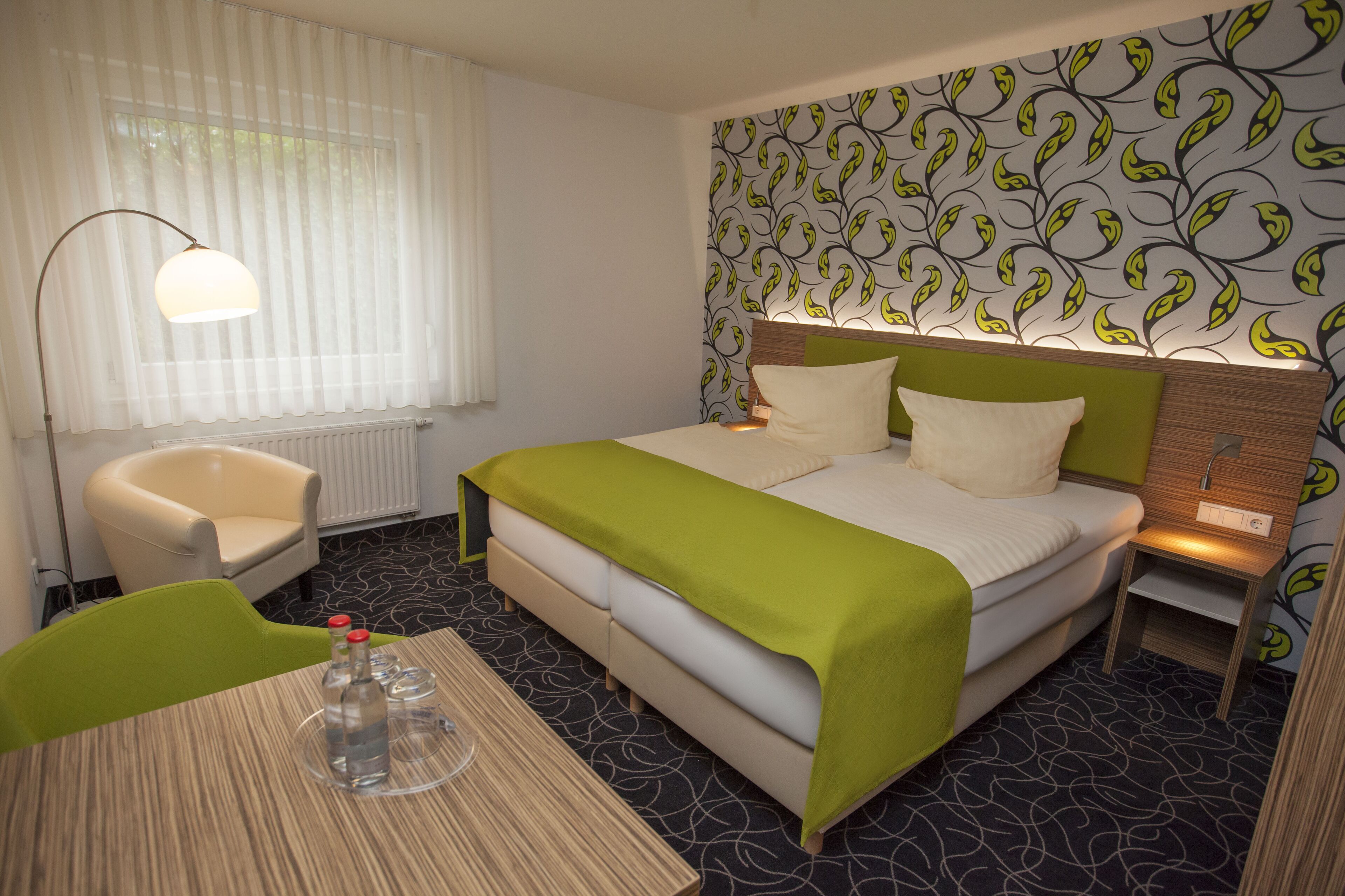 A hotel room with a bed, a chair, a table with bottles, and a patterned wall.