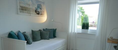 Una habitación con cama blanca, almohadas verdes, radiador blanco, ventana con vistas a follaje y un escritorio con silla.