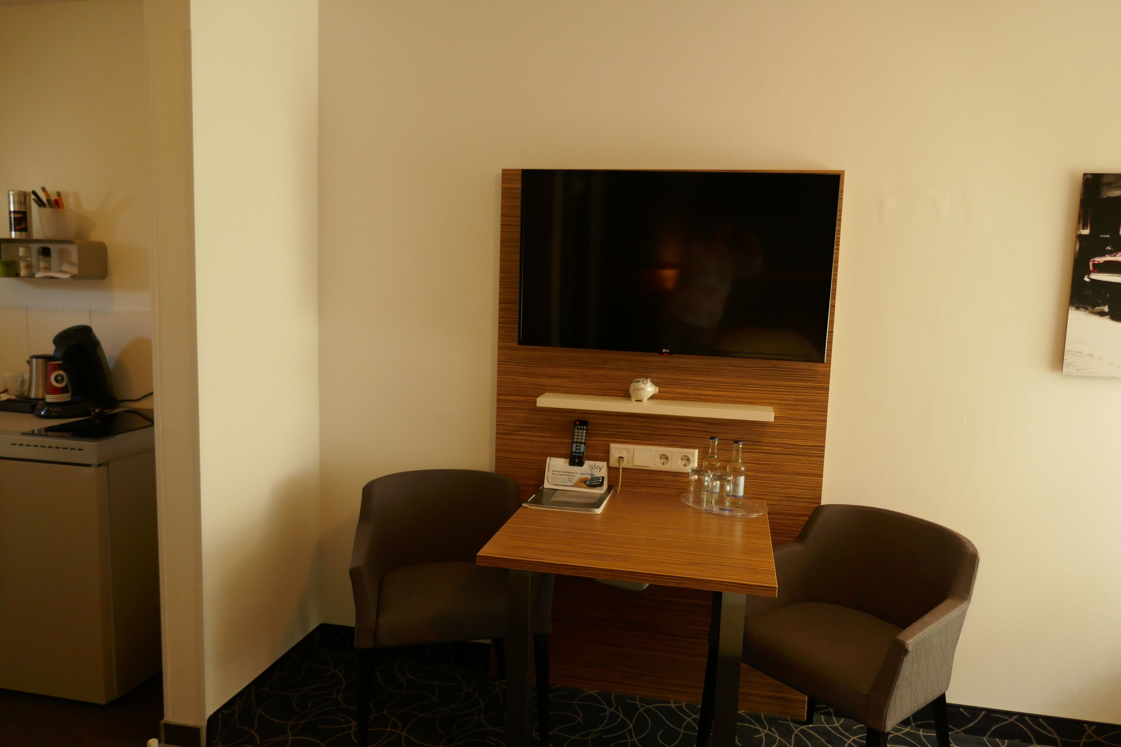 Ett hotellrum med ett träbord, två stolar, en platt-TV som är monterad på väggen, en liten hylla med en fjärrkontroll och en kaffemaskin.