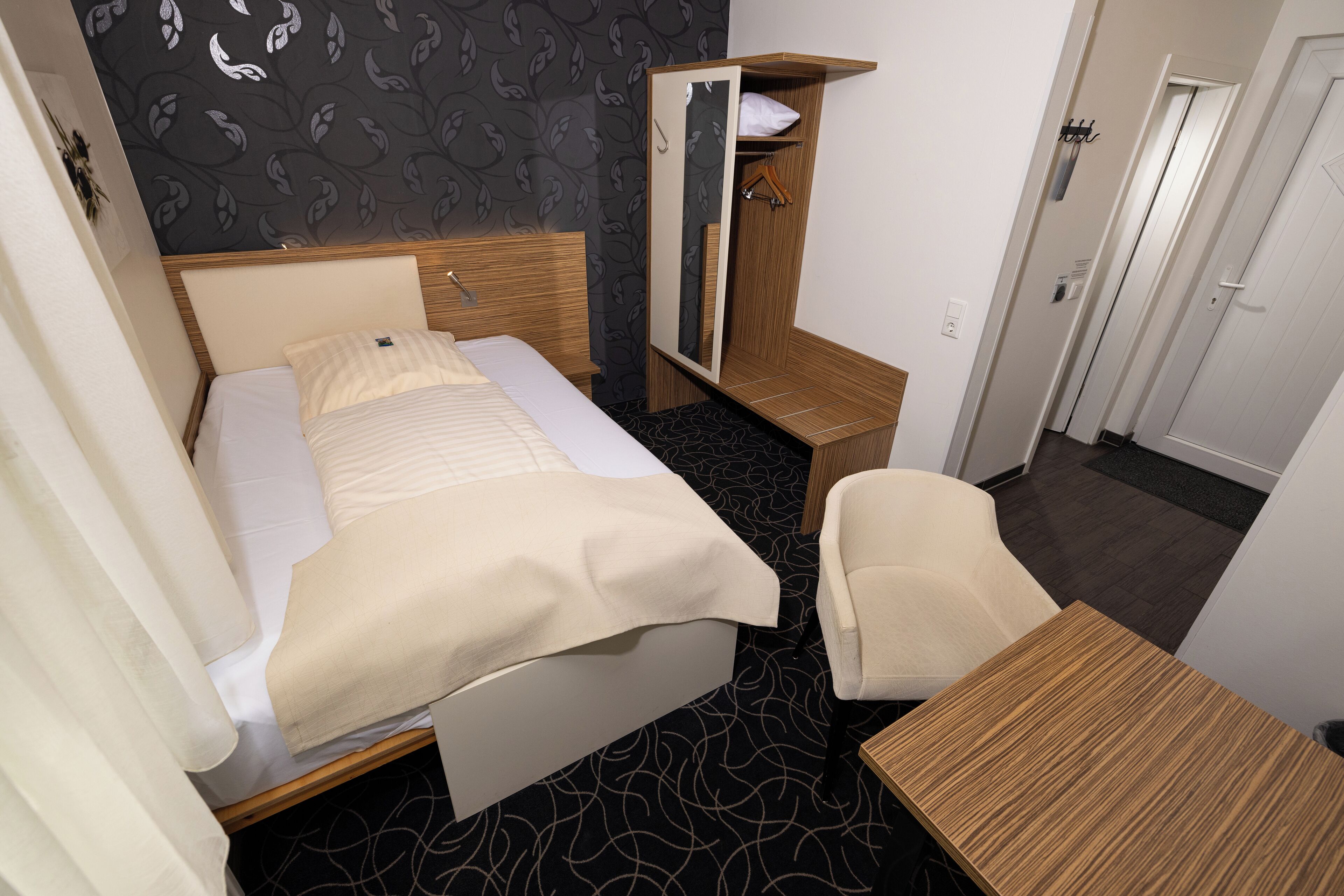 A neatly arranged hotel room with a bed, a wooden nightstand, a chair, a wardrobe, and a small table.