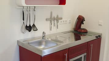Eine kleine Küche mit Spüle, Kaffeemaschine und Mikrowelle. Küchenutensilien hängen an der Wand und ein kleines Topfpflänzchen steht auf einem Regal.