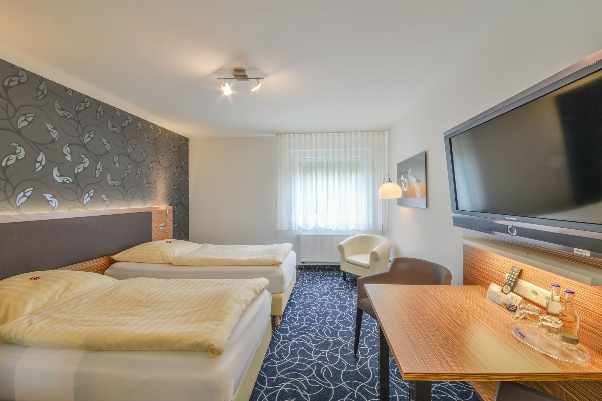 A hotel room with two beds, a TV, a desk with a tray of toiletries, a chair, and a window with sheer curtains.