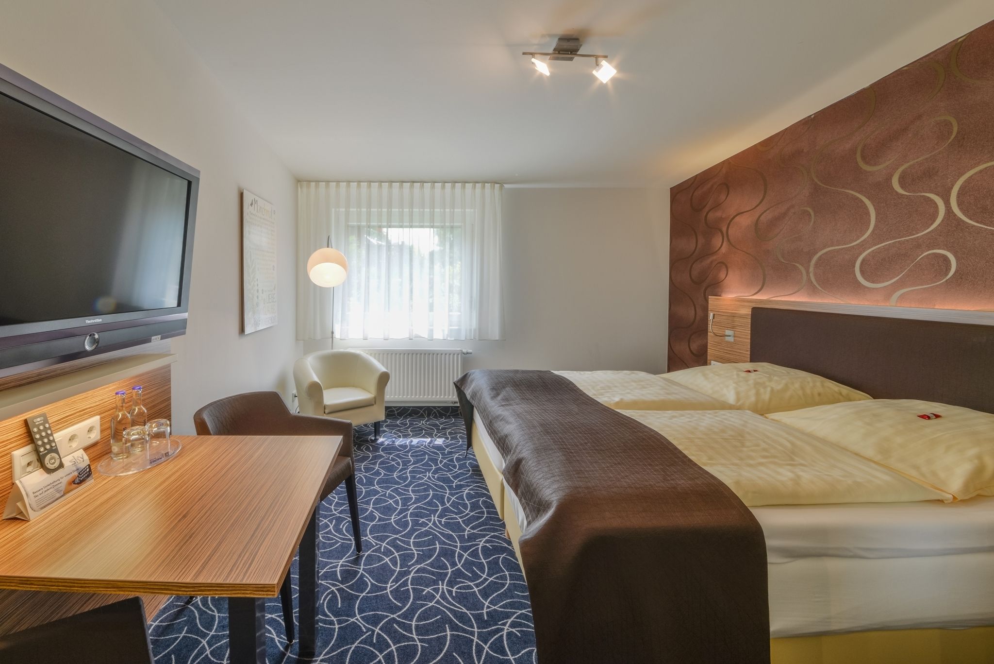 A hotel room with a bed, a desk with a flat-screen TV, a chair, a table with a remote control, and a window with curtains.