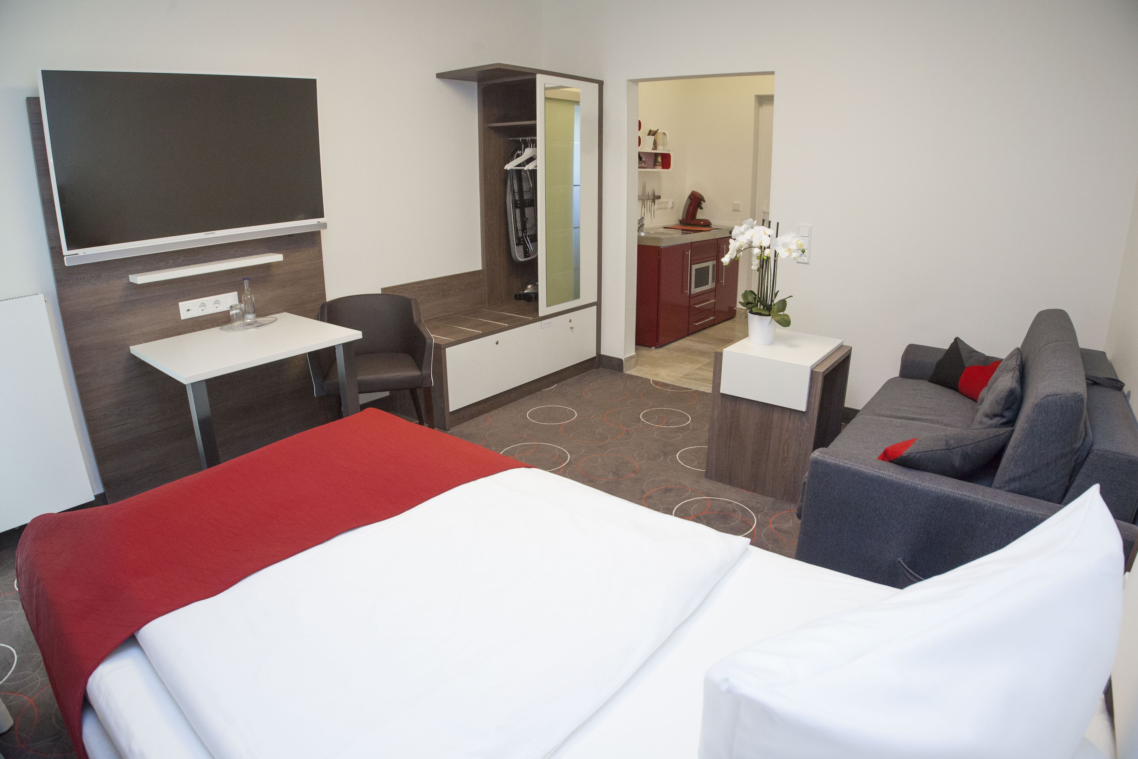 A hotel room with a bed, a desk, a sofa, a TV, a mirror, and a bathroom visible through an open door.