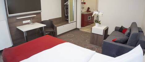 Habitación de hotel con cama, escritorio, sofá, televisor, espejo y baño visible a través de una puerta abierta.