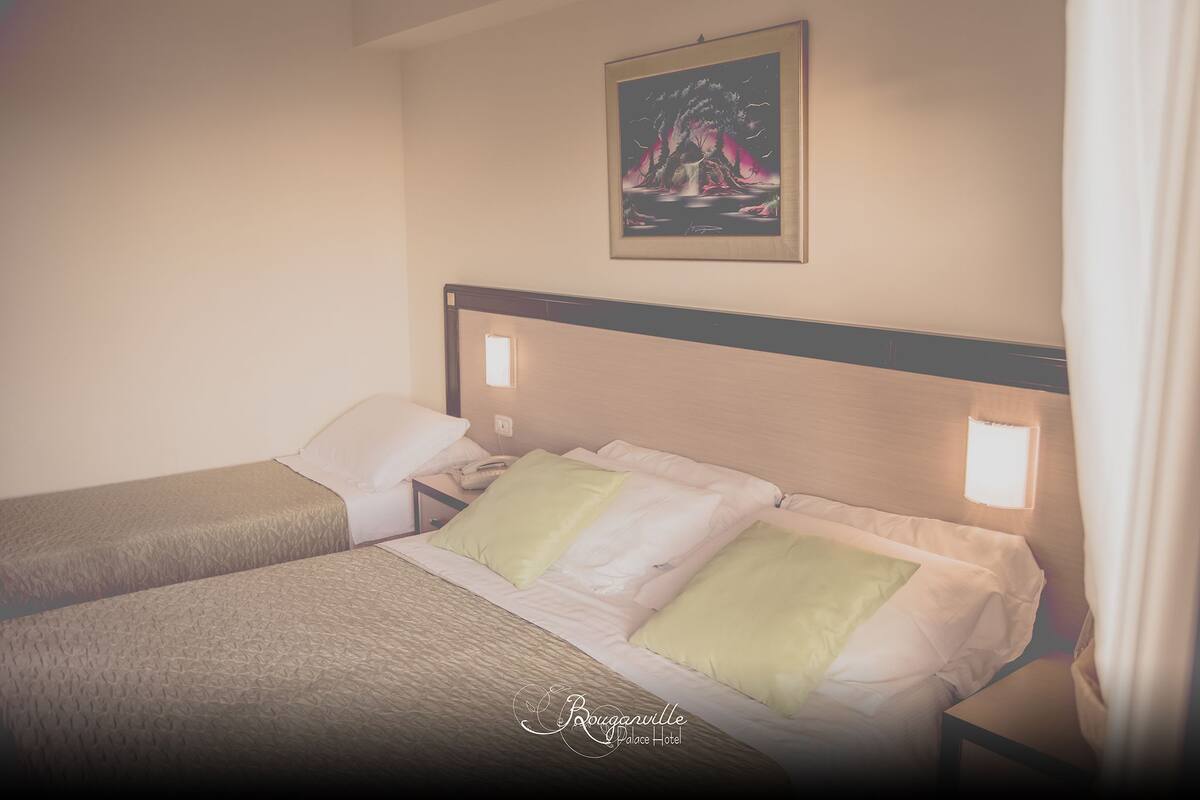 A hotel room with two beds, a nightstand, a painting on the wall, and a window with curtains.