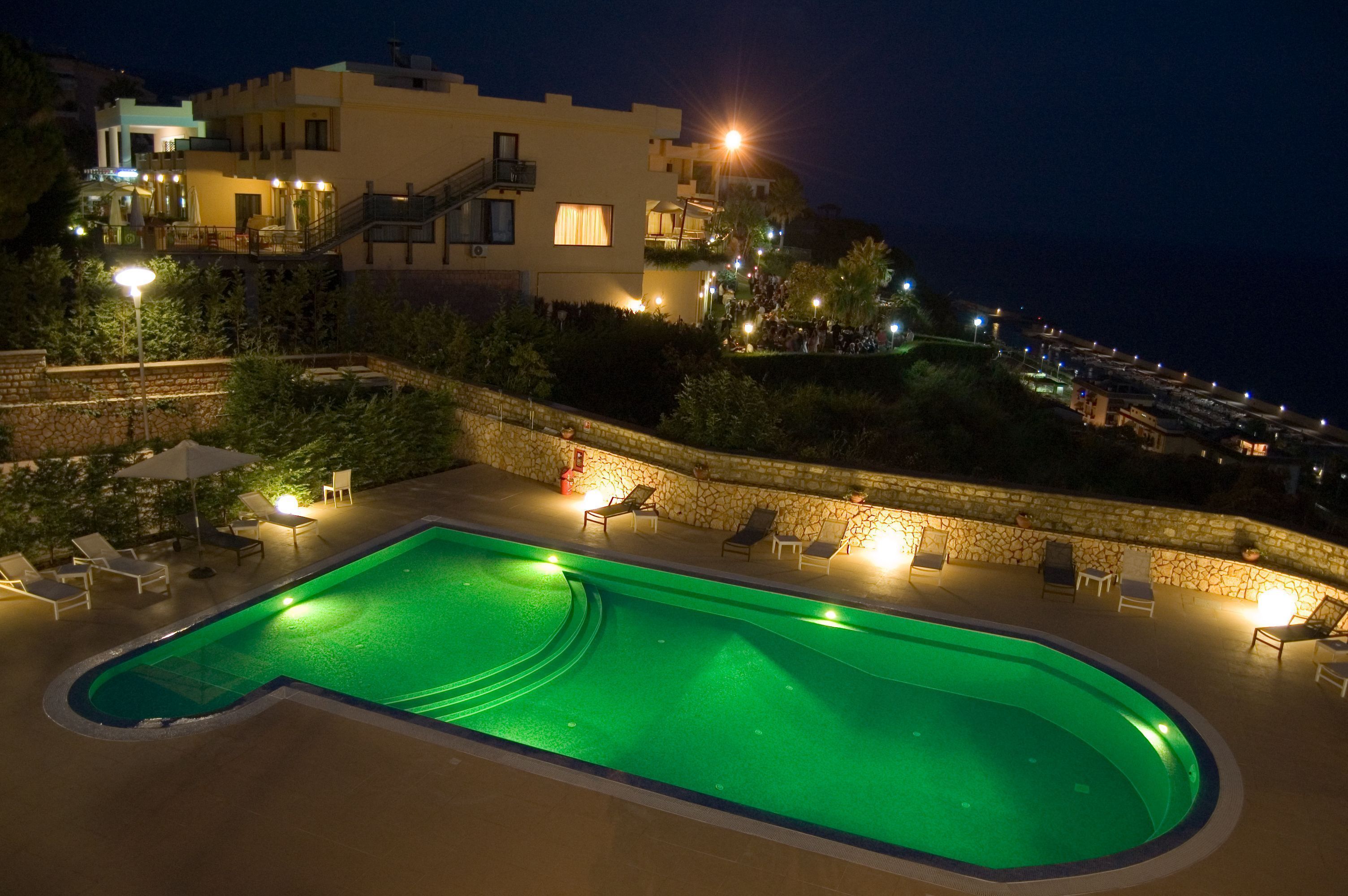 Una piscina con sillones de descanso y un muro de piedra, rodeada de edificios con luces.