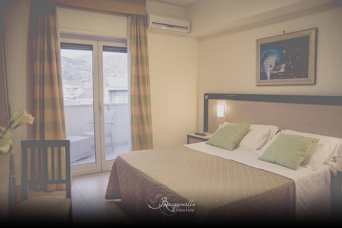A hotel room with a bed, a chair, a nightstand, a painting on the wall, and a view through the window.