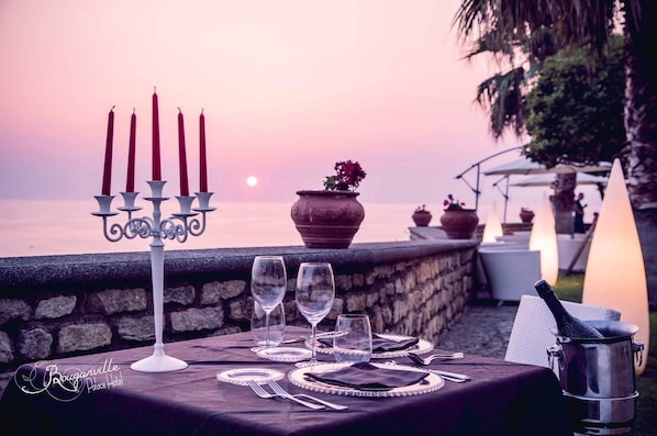 A table set for a meal with candles, wine glasses, and a bottle of champagne, overlooking a sunset over the water.