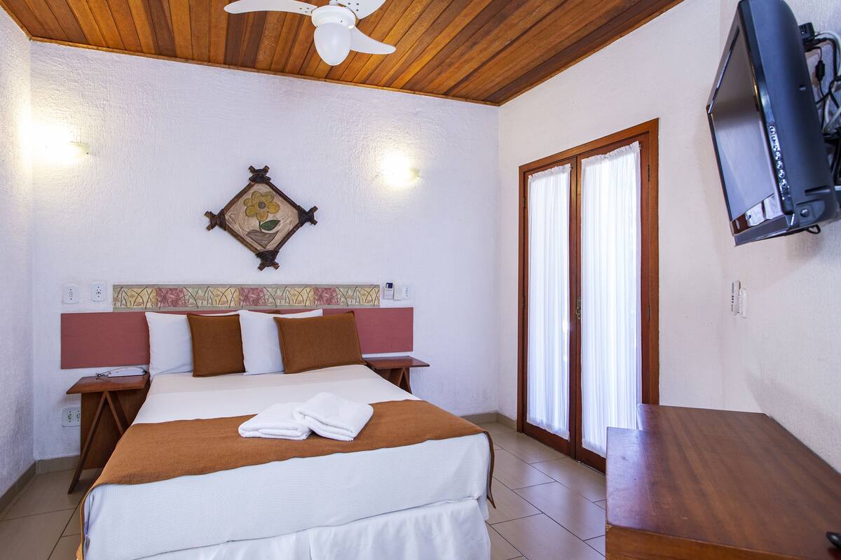 A hotel room with a bed, wooden headboard, bedside tables, a television, and a window with curtains.