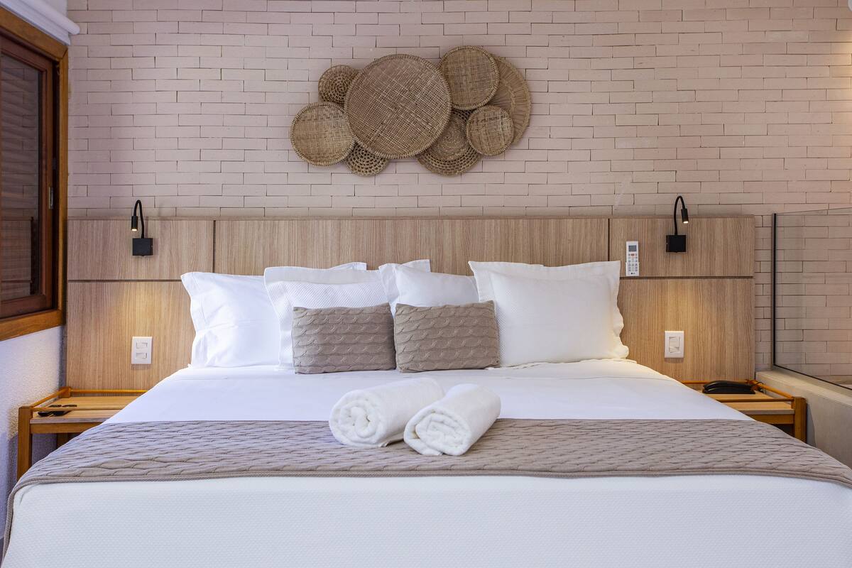 A neatly made bed with white linens, two rolled towels, and a woven wall decoration above it.