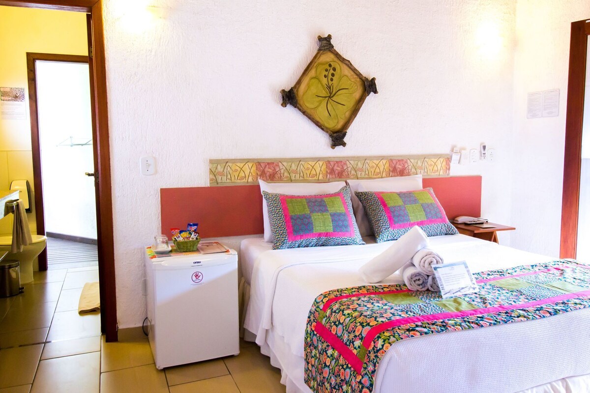 Une chambre d’hôtel avec un lit, deux tables de chevet, une petite table, une horloge murale et une fenêtre.