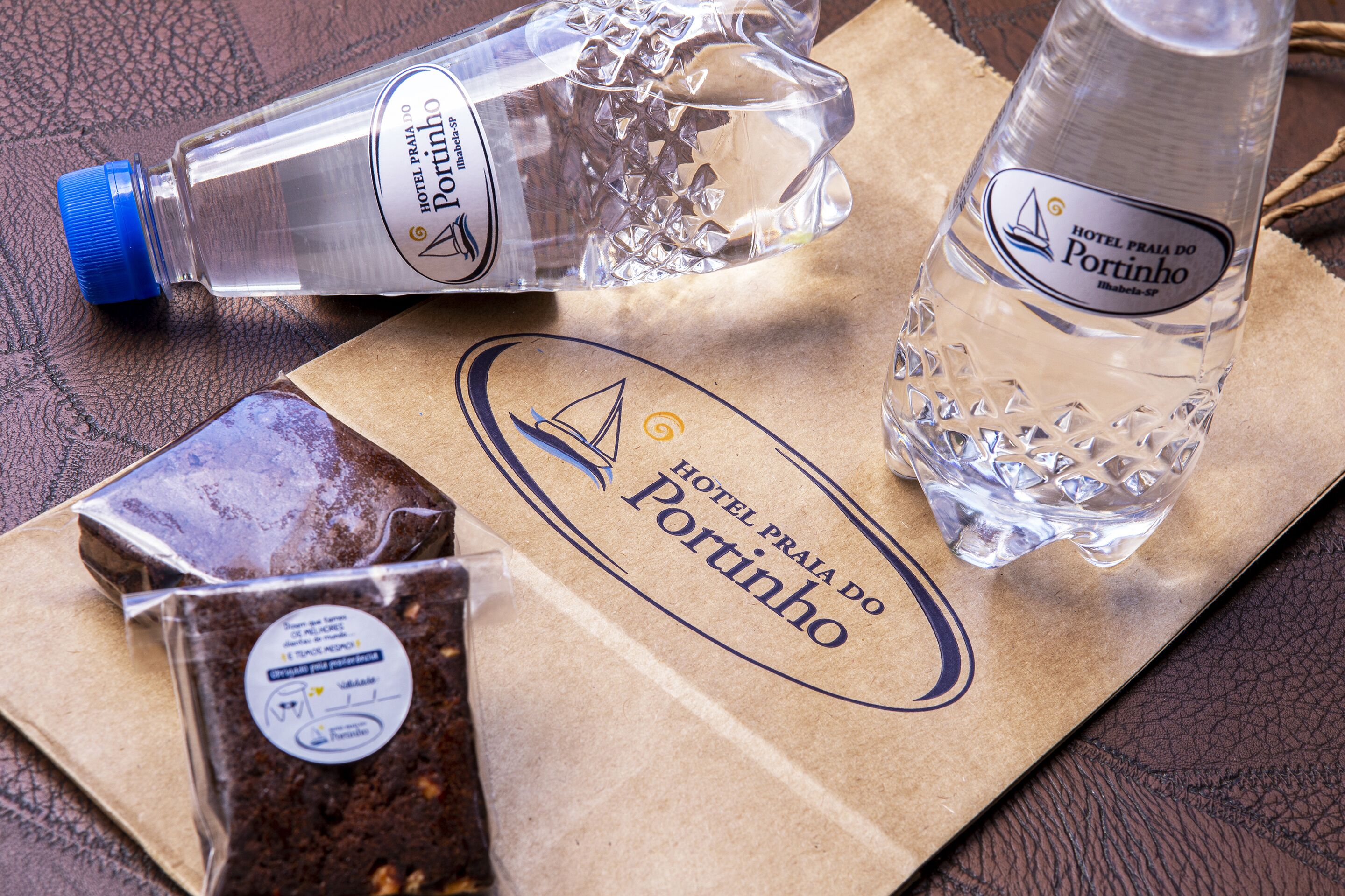 Een bruine papieren tas met het logo van Hotel Praia do Portinho, met daarin een waterfles en een chocoladereep.