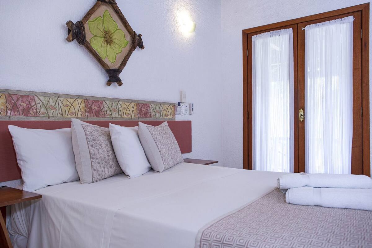 A bedroom with a bed, pillows, a nightstand, a wall clock, and a window with curtains.