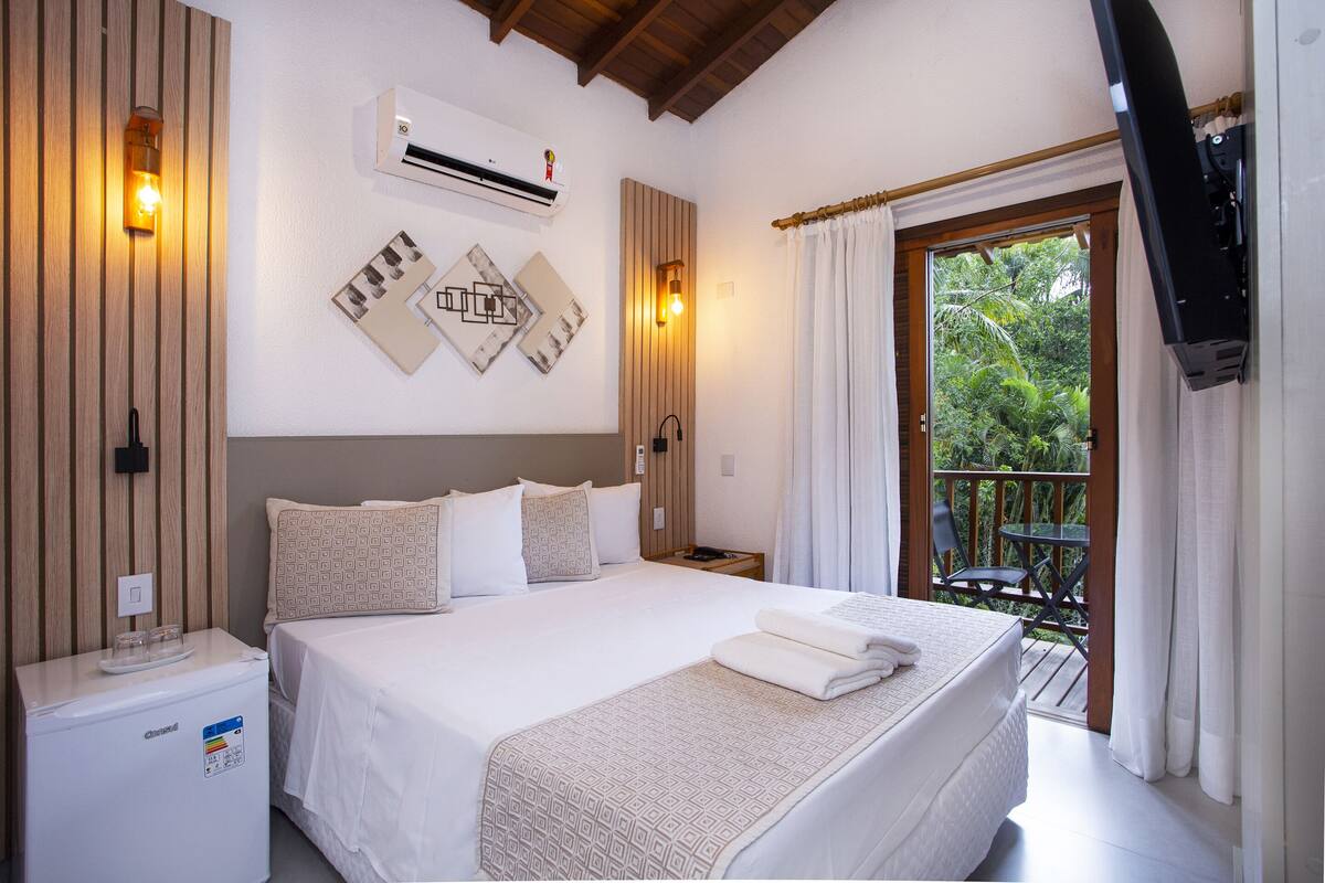 A hotel room with a bed, a small table, a chair, an air conditioning unit, and a balcony with a view of greenery.