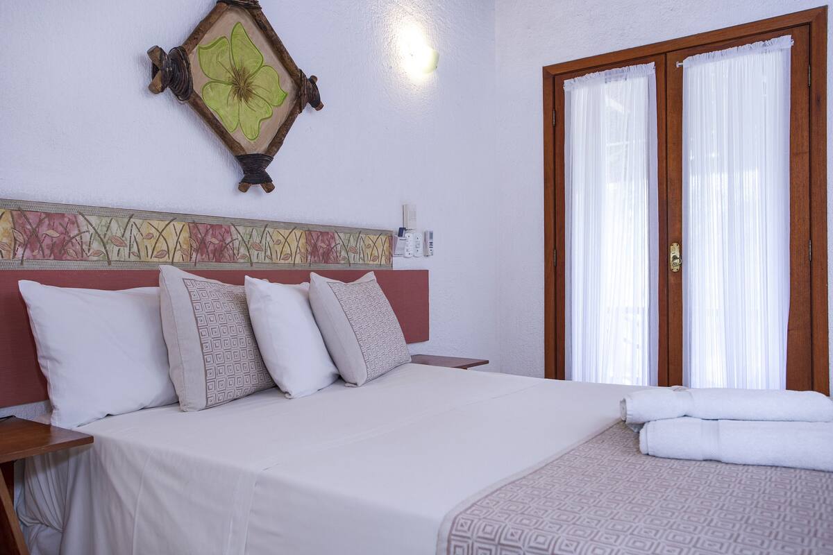 A neatly made bed with white linens and patterned pillows, a wooden headboard with a floral design, and a window with white curtains.
