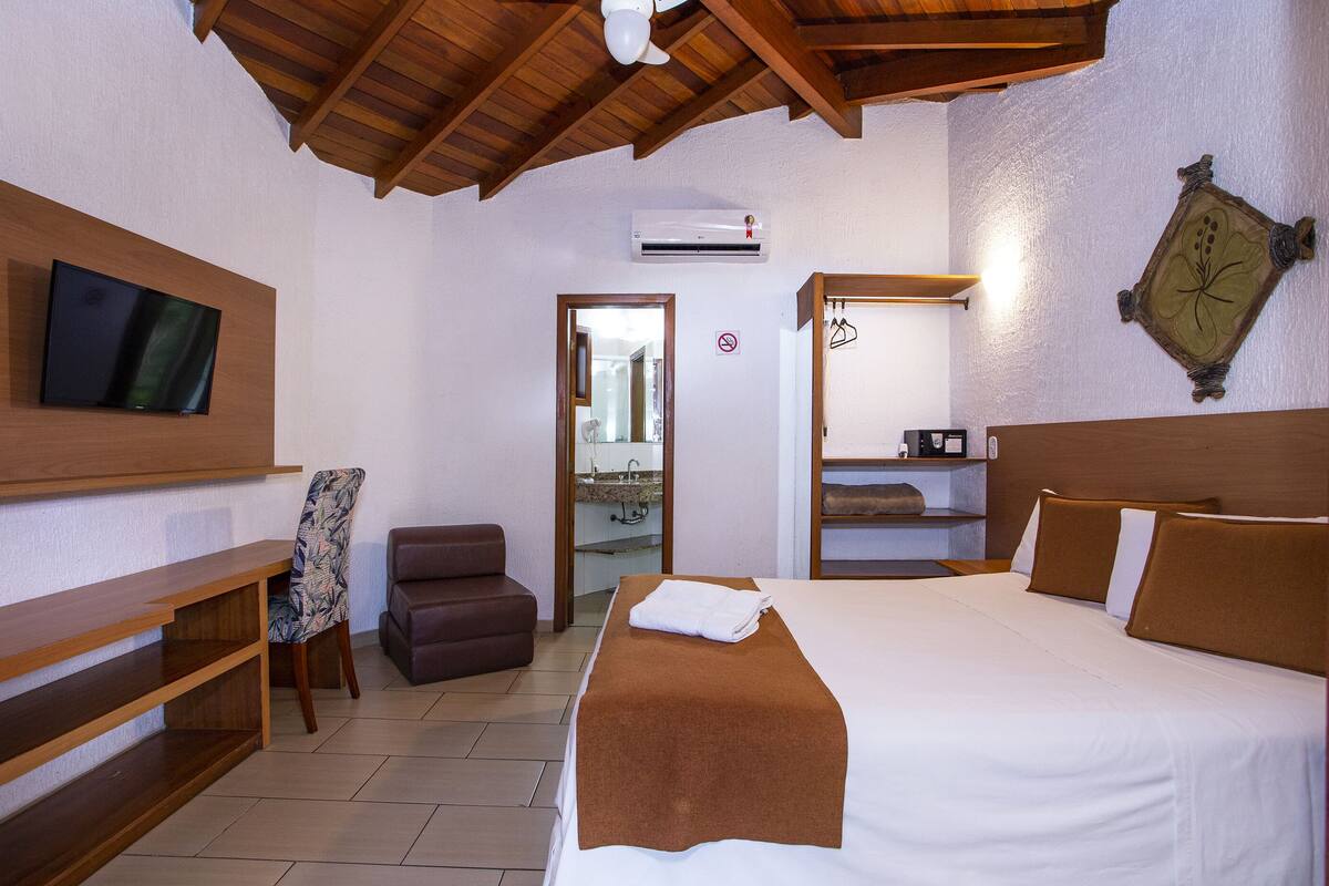 A hotel room with a bed, a TV mounted on the wall, a wooden bench, a chair, and a small table.